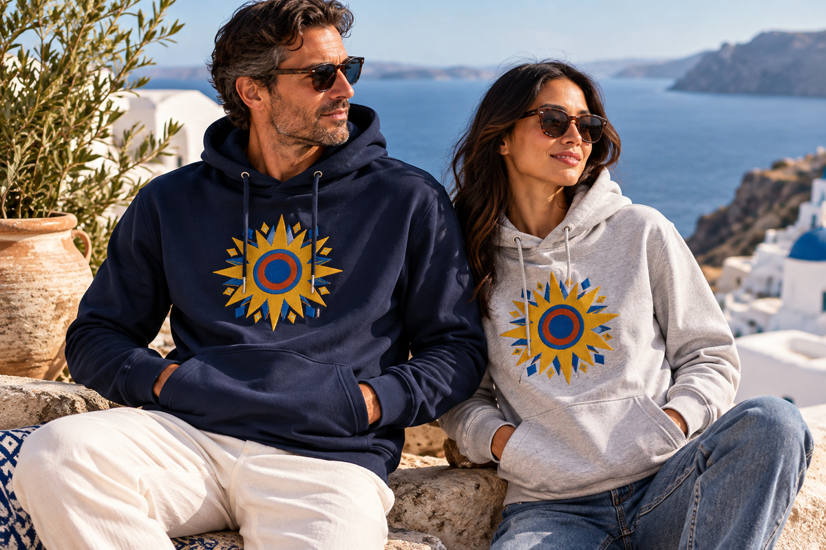 Male and female models wearing Asteri Solaris embroidered unisex hoodies in French Navy and Heather Grey, featuring colorful Mediterranean sunburst designs on a sunlit Greek island terrace.